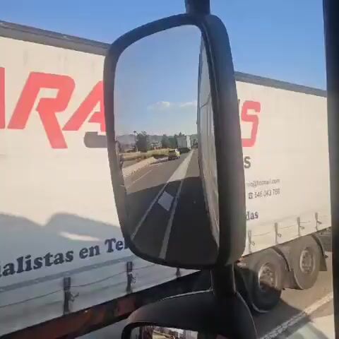 The video is filmed from the perspective of a truck driver. A side mirror reflects the highway ahead, showing a line of trucks and cars. The highway has a concrete barrier with yellow reflectors. The surrounding environment appears to be dry and hilly, with some scattered trees. In the background, there is smoke rising from an area, possibly a fire. The sky is clear and blue with a few white clouds.