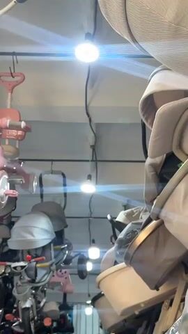 The video shows an indoor marketplace or store where children's items are displayed. The primary focus is on several pink and white children's tricycles and ride-on toys hanging from the ceiling or mounted on display racks. In the background, there are also what appear to be baby strollers and cribs. The lighting is bright, with overhead lights illuminating the space. The scene is densely packed with merchandise, suggesting a retail environment.