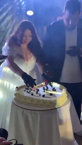 A woman in a white dress and a man in a black suit are cutting a heart-shaped cake. The cake is topped with berries. The woman is smiling and the man is focused on cutting the cake. They are both wearing black gloves. The background appears to be a dimly lit event space with stage lighting.
