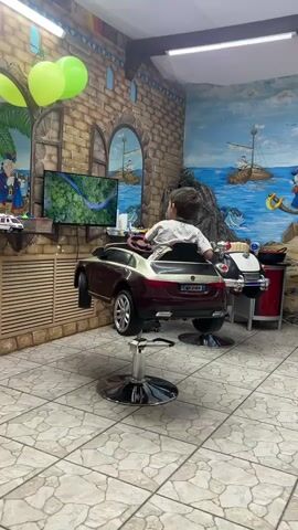 A young boy is sitting in a toy car that is mounted on a barber chair. The boy is wearing a white t-shirt and is looking at a screen that is displaying a nature scene. The background of the scene is a mural of a coastal town with boats on the water. There are also balloons and other decorations in the room. The boy appears to be interacting with the screen, possibly playing a game or watching a video. The room looks like a children's hair salon.