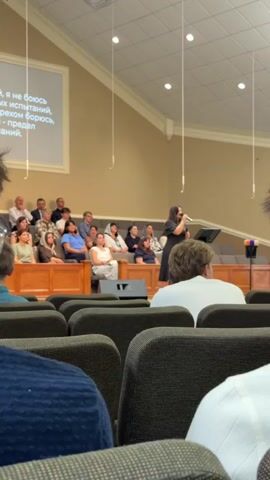 A woman is singing at a podium in front of a congregation. The woman is wearing a black dress and is holding a microphone. The congregation is seated in rows of chairs in front of the woman. There is a screen behind the woman with text in Russian. The room is well-lit and appears to be a church or other place of worship. The video is taken from behind the congregation, looking towards the stage. The focus is on the woman singing, with the congregation slightly blurred in the background.