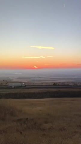 A static, eye-level shot captures a wide landscape during sunset. The sky transitions from a light blue at the top to hues of orange and red near the horizon. A few thin, elongated clouds are visible in the upper part of the sky, with a faint white streak, possibly a contrail, high above. The sun is partially visible just above the horizon, appearing as a bright, hazy orb with a few dimmer segments.

In the middle ground, a highway runs horizontally across the frame. On the highway, a white semi-trailer truck is traveling from left to right, with a small white car positioned behind it and further to the left. The truck appears to be the primary object of focus in this middle section.

The foreground consists of a grassy hill covered in dry, golden-brown grass, sloping downwards towards the highway. The terrain beyond the highway recedes into the distance, appearing as a flat, hazy expanse with subtle variations in color, suggesting agricultural fields or plains. No people are directly visible. The overall impression is one of a serene, natural landscape at dusk with elements of human infrastructure present.