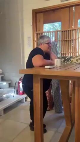 A woman is standing at a wooden table in an indoor space. She is leaning against the table, with her left arm resting on the surface and her head resting on her hand. She is wearing a dark blue shirt, black leggings, and black sneakers. She has short, gray hair and wears glasses. On the table, there is a white mug and a small white object that might be a remote. The woman is looking to her right and appears to be listening or thinking. Behind her, there's a door with wooden slats, and two rolls of fabric with different patterns are leaning against the door. To her left, there is a white staircase with a red fire extinguisher positioned near it. The walls are painted white with vertical wood panels. The floor is tiled in a light color. Someone off-camera says "Basta." Then she says, "Ah, mira. ¿Que vas a hacer?" which means "Oh, look. What are you going to do?" in English. Someone off-camera says, "Pasta."