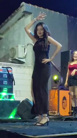 The video shows a woman in a black dress dancing on a stage. She has long, dark hair and is wearing high heels. The stage appears to be outdoors, with a building and a roof in the background. There are also speakers and lighting equipment on the stage. Another woman in a red top and black skirt is visible to the right of the frame. The woman in the black dress is moving her arms and body in a fluid, graceful manner, seemingly to music. Her right hand is on her head, and her left hand is on her hip. The dress has a thigh-high slit. There is a slight smile on her face. The lighting is dim, with some purple and orange lights illuminating the stage.