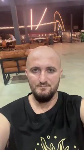 A bald man with a beard, wearing a black t-shirt, is looking at the camera in what appears to be an outdoor seating area at night. He is seated in a chair with brown cushions visible. In the background, there are tables and chairs, string lights overhead, and a large illuminated structure that resembles a roller coaster or Ferris wheel. Some other people are seated at tables in the background, out of focus.