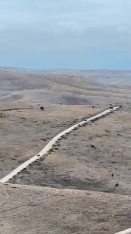 An aerial drone shot captures a desolate landscape with rolling hills under a cloudy sky. A long, dusty road winds through the center of the frame, with several vehicles visible on it. The terrain is dry and covered in sparse, brown vegetation, with occasional small trees scattered throughout. The scene conveys a sense of remoteness and isolation.