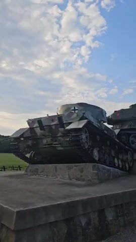 A low-angle shot shows two tanks on display outside. The tank in the foreground is camouflaged and appears to be a German tank with a German cross on its turret. The tank behind it is green and has a red star on its turret, indicating it is a Soviet tank. A young boy in an orange shirt and yellow shorts stands on the right side of the image, next to the Soviet tank. He is facing the tanks and pointing towards the Soviet tank. The tanks are mounted on stone pedestals. The sky is blue with scattered clouds.