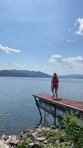 A young person in a red swimsuit runs to the edge of a wooden dock and jumps into the water. The dock is supported by large, gray pontoons. The water is a deep blue and calm, with a few small whitecaps. In the background, rolling green hills meet a clear blue sky with a few fluffy white clouds. The scene is bright and sunny.