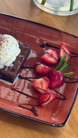A close-up shot of a dessert plate on a wooden table. The plate is rectangular with a reddish-brown speckled glaze. On the plate is a square chocolate brownie topped with a scoop of vanilla ice cream dusted with cocoa powder. Melted white cream surrounds the brownie. To the right of the brownie are several sliced strawberries and a sprig of mint. Chocolate syrup is drizzled across the plate. The video is a static shot, with no discernible action or people visible.