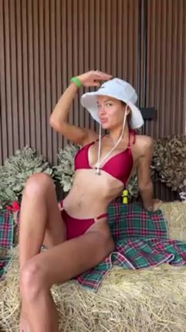 A woman is sitting on a bale of hay. She is wearing a red bikini and a white sun hat. The bale of hay is covered in a red and green plaid blanket. Behind the woman is a brown wall and some dried plants. The woman looks up and stretches her arms out to the side.