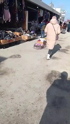 The video shows a person walking on a paved path in what appears to be an outdoor market. The path has patches of light and dark coloring. On the left side of the path are market stalls with various items for sale, including footwear, clothing, and packaged goods.  Several people are visible near the stalls, some appearing to be browsing or shopping.  To the right of the path, more people are walking in the same direction as the person filming. The sky is clear and blue. The shadow of the person filming stretches out onto the path.
