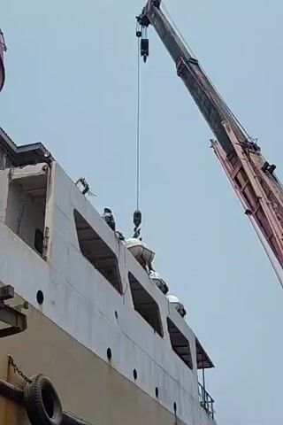 A crane hoists a life raft from the deck of a ship. Two workers, wearing hard hats and safety vests, stand on the deck, one of them near the life raft. The crane's boom is visible at the top right of the frame. The ship appears to be in port or undergoing maintenance. The sky is overcast.