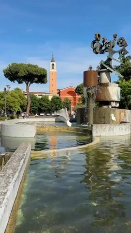 The video shows a fountain with a light green pool of water surrounded by low gray concrete walls. A metal sculpture is on one side of the fountain, and a few gulls are visible around it. In the background, there are trees and a tall church with a bell tower. People are sitting on benches in the square. The water in the fountain is flowing. The sky is blue with a few clouds.