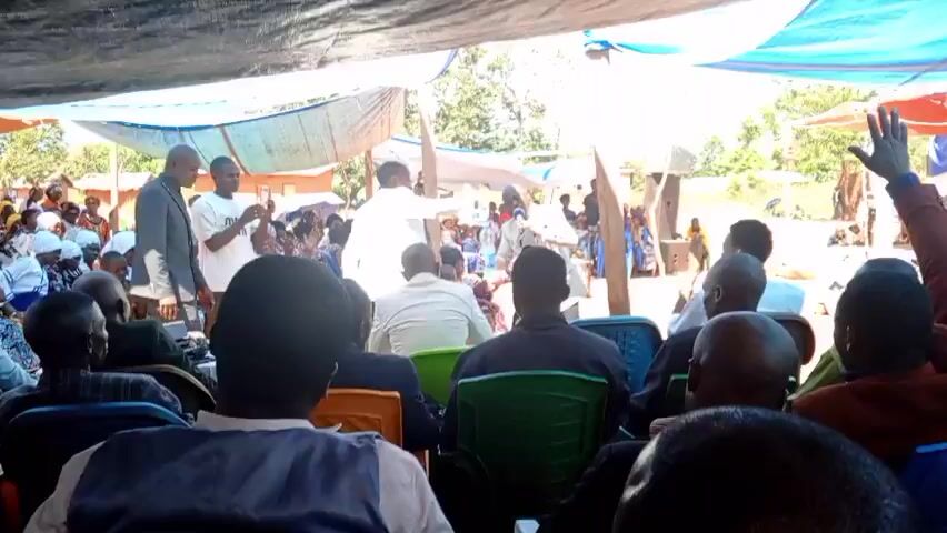 Under a makeshift tent structure, an outdoor gathering takes place. A man in a light-colored suit holds a microphone and water bottle, speaking to a crowd of seated individuals, many of whom are facing him. Another man in a white suit stands beside the speaker, gesturing with his hand. People in the crowd are recording with their phones. Trees and a building can be seen in the background.