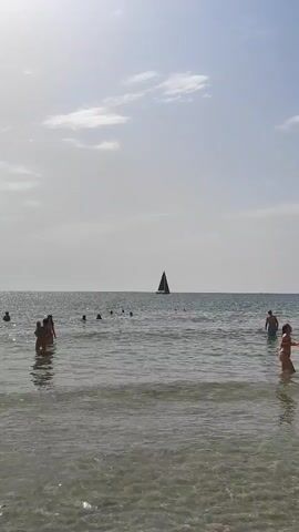 The video shows a body of water with a clear sky. In the distance, a sailboat with a dark sail is visible. Several people are in the water, some appearing to be swimming or wading. The water is calm, with small waves. The overall scene is one of a beach or coastal area on a clear day.
