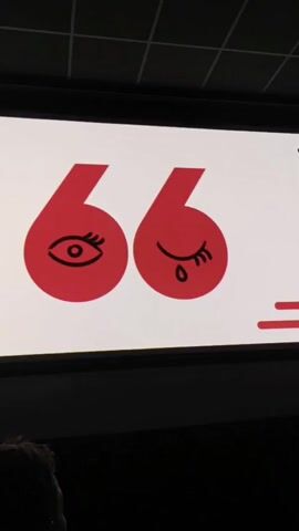 The video begins with a screen showing the logo for the Zlín Film Festival, featuring two red shapes resembling the number “6” with stylized eyes, one open and one with a teardrop. The camera then moves back to show a dimly lit theater with rows of seats.  People are seated in the audience.  In the foreground, a doll dressed as a nun is sitting upright in a seat, making a “rock on” hand gesture with both hands. Other members of the audience are visible in the background.