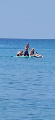Three people are on a paddleboard in the ocean. The person in the back is standing and holding a paddle, and appears to be paddling. The other two people are sitting on the paddleboard. One is leaning forward, and the other is lying down. The water is blue, and the sky is clear. A yellow inflatable tube is visible in the distance, being pulled by a boat.