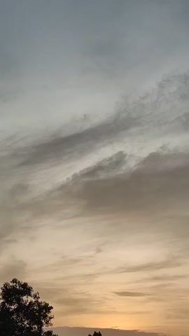 The video shows a time lapse of a sunset over a city skyline. The sun is visible as a bright orange orb peeking through a layer of clouds. The sky transitions from a pale yellow to a light orange as the sun sets. Buildings and trees are silhouetted against the colorful sky.