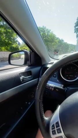 The video shows a view from inside a car, looking out the side window and side mirror. The sun is shining brightly, casting a glare on the windshield and creating lens flare. Trees and a wall are visible outside the car. In the side mirror, another vehicle, possibly a motorcycle, is seen driving on the road in front of the car. The interior of the car is visible, including the door panel, window controls, and part of the steering wheel. The road appears to be paved and is bordered by greenery. The overall impression is of a sunny day drive.