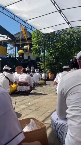 The video shows a group of people sitting on the ground outdoors, facing a temple-like structure. They are all dressed in white, traditional clothing. A young girl in the foreground is wearing a white outfit and raises her middle finger towards the camera. The setting appears to be a cultural or religious event.