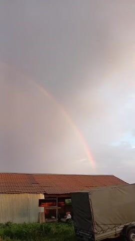 The video shows a slightly overcast sky with a faint rainbow arching across it. Below the sky, there are several buildings with red tiled roofs and corrugated metal siding. There are also some plants growing around the buildings, and a small truck is partially visible on the right side of the frame. The scene appears to be a rural or semi-rural area. The video is taken from a static position, and there are no visible people or significant actions taking place. The lighting suggests it is either early morning or late evening.