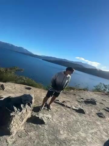The video shows a young man standing on a rocky cliff overlooking a vast body of water. He is wearing a gradient gray and black Nike hoodie, dark shorts, and sandals, and is sporting sunglasses. The camera then shifts to showcase the landscape, revealing a serene lake surrounded by tree-covered mountains under a clear blue sky. The water is calm, with gentle ripples creating a subtle texture on the surface. The environment suggests a peaceful, natural setting.