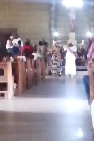 The video shows the interior of what appears to be a church. People are seated in wooden pews on either side of an aisle. In the distance, towards the front of the church, there is a group of people, including a person in a long white dress with a white sash. Above them, a statue of a person is on a cross. The church has a patterned wallpaper. The lighting is bright, but the image is somewhat blurry. There is singing audible in the background.