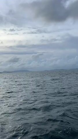 A slow pan reveals an overcast sky with scattered clouds above a choppy, dark blue ocean. In the distance, a faint, hazy line of islands or a coastline is visible. The camera then pans slightly to the right, showing the edge of a white boat with a pink cushion in the foreground. The sun is partially visible through a break in the clouds, casting a soft glow on the water.