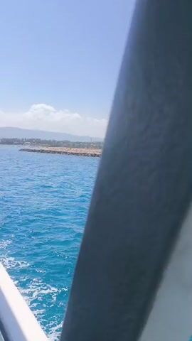 The video pans across a bright blue ocean with gentle waves. In the background, a coastline is visible with buildings and a rocky breakwater. The sky is clear with a few scattered white clouds. The camera appears to be moving, possibly from a boat.