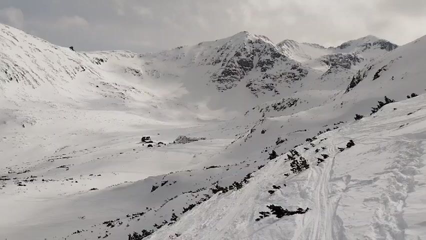 The video shows a snowy mountain landscape. A few buildings are visible in the valley below. There are tracks in the snow, possibly made by skis or other equipment. The sky is overcast.