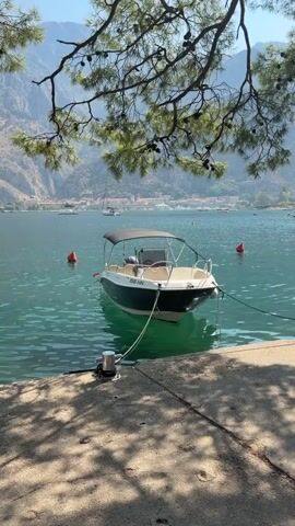 A small motorboat is docked on the clear, turquoise water. The boat is white and dark green, with a black canopy and a license plate that reads "156 HN." It is tied to a metal cleat on a concrete pier. In the background, there are mountains, a town with red-tiled roofs, and other boats, including sailboats. The foreground is shaded by the branches of a pine tree. The sun is shining, and the sky is blue.
