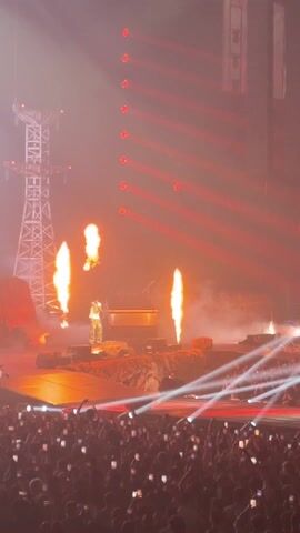 A medium shot captures a performer on stage amidst a concert setting. The stage is set up with pyrotechnics, featuring flame bursts and a rocky terrain. The performer, dressed in light-colored attire, stands center stage, illuminated by bright spotlights. The backdrop is enhanced with a tower-like structure on the left and vertical rows of red lights on the right. A dense crowd of people is visible in the foreground, many holding up their phones to record the performance. The atmosphere is vibrant with a combination of white and red lighting effects.