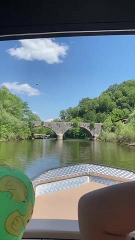 The video shows a view from a boat moving down a river towards a stone bridge with multiple arches. The bridge is old and covered in greenery, suggesting it is situated in a natural, possibly historical, setting. Lush green trees and hills line the banks of the river, indicating a temperate climate and a scenic, rural location. The water of the river is calm and reflects the bright blue sky, which has a few scattered white clouds. The scene appears to be set during the day, likely late morning or early afternoon, given the brightness of the sunlight.

In the foreground, the front of the boat is visible, with a metallic textured surface and a tan-colored seat. A person's arm, possibly wearing a green garment with a cartoon elephant pattern, is visible on the left side of the frame, suggesting the presence of at least one person on the boat. The overall mood conveyed is peaceful and serene, with the gentle movement of the boat and the natural beauty of the surroundings creating a relaxing atmosphere. The bridge in the background serves as a focal point, hinting at a journey towards a destination or a point of interest. The presence of other boats visible under the bridge suggests that this is a popular spot for recreational activities on the river. The location appears to be Rijeka Crnojevića in Montenegro, given the characteristic stone bridge and surrounding landscape. There is no visible text in the video.