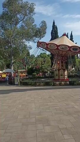 This video captures a swing ride in an amusement park, likely in Bolshoy Sochi, Russia. The ride itself is a classic carousel-style swing ride with a prominent red and cream-colored canopy adorned with decorative elements. The central column of the ride is elaborately decorated with panels featuring what appear to be scenic paintings or designs, possibly depicting various historical or fantastical scenes. Attached to the canopy are numerous swings, each with yellow seats and metal chains, suspended from the main structure.

Several people are visible on the swings, enjoying the ride. They appear as small figures against the backdrop of the park. The atmosphere suggests a pleasant, possibly sunny day, with a bright blue sky and scattered clouds. Lush green trees and foliage surround the ride, indicating a well-maintained park setting. The ground in the foreground is paved with large, light-colored stone tiles, suggesting a public walkway or plaza area within the park.

The ride is in motion, with the swings arcing outwards, indicating that the attraction is operational. The overall scene conveys a sense of leisure and entertainment. The presence of tall, slender evergreen trees in the background adds a distinctive visual element that, combined with the overall greenery and the style of the amusement park, aligns with the provided location context of Bolshoy Sochi. The time of day appears to be daytime, given the bright lighting and shadows. No discernible text is visible on the ride or in the immediate surroundings.