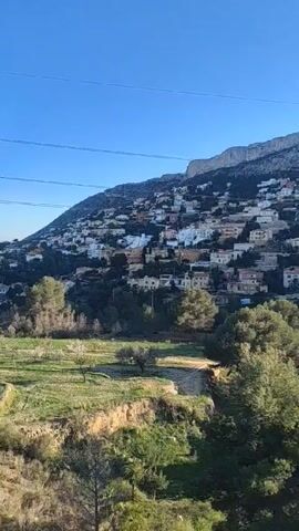 The video shows a hillside community of houses on a sunny day. The houses are predominantly white and are scattered across the hillside, blending with the natural vegetation. A clear blue sky is visible above, punctuated by power lines. In the foreground, a green field stretches out with scattered trees. A road is visible in the very foreground. The general atmosphere is peaceful and idyllic.