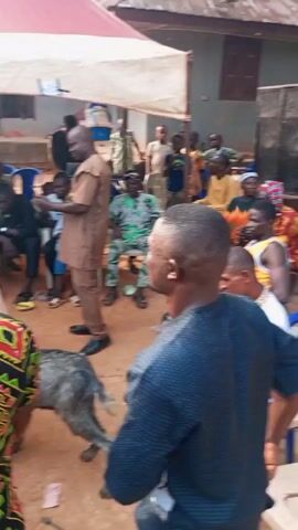 The video depicts a lively outdoor gathering under a white and red tent. Numerous people of various ages and attire are present, including some wearing traditional African prints. A man in a brown outfit stands near the front left, seemingly observing or gesturing. A grey goat wanders freely amidst the crowd. Individuals are conversing, moving around, and some are dancing to music. The setting appears to be a village or rural area, with houses visible in the background. The atmosphere suggests a celebration or community event.