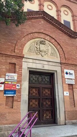 A frontal medium shot of a building facade shows an arched entryway with a large wooden door and a decorative crest above it. The building is constructed of red brick. A sign with the letters "FEC" and a logo is visible on the right side of the door. Another sign with "Erasmus+" and other logos is on the left. A set of purple railings leads up to the entrance. The number 25 is visible above the door. The overall scene is bright and appears to be daytime.