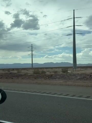 The media file captures a drive through a semi-arid landscape, likely in or near Boulder City, USA. The scene is dominated by a vast, open sky with dramatic, heavy clouds, suggesting overcast weather or the approach of a storm. Towering electrical pylons, characteristic of power transmission infrastructure, are a prominent feature, stretching across the horizon and marking the passage of the highway.

The viewpoint is from inside a vehicle, looking out through the windshield and side window. The road ahead curves gently, with other vehicles visible in the distance, indicating ongoing travel. The terrain is rugged and dry, with sparse vegetation and rocky ground. In the far distance, a range of mountains forms a hazy backdrop. The side mirror of the car is visible, reflecting a portion of the scene, and displays the text "OBJECTS IN MIRROR ARE CLOSER THAN THEY APPEAR."

The overall mood is one of a journey through a somewhat stark, industrial-tinged natural environment. The time of day is difficult to pinpoint precisely due to the cloud cover, but it appears to be daytime. The activity is simply driving, with the camera capturing the passing scenery. There are no people clearly visible as subjects within the frame, though their presence is implied by the vehicle and the journey itself. The setting evokes a sense of scale with the vastness of the sky and land, contrasted with the man-made structures of power lines and the highway.