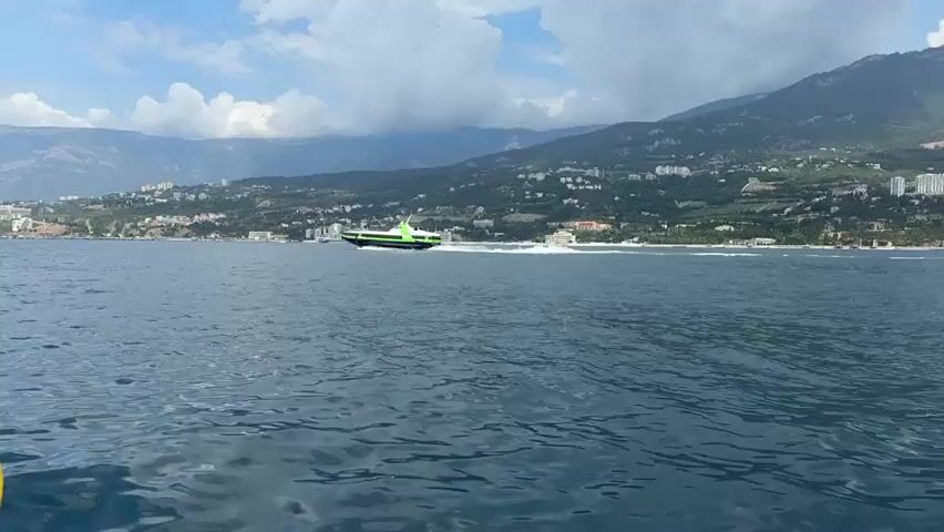 A green and black hydrofoil is speeding across the water, leaving a white wake behind it. The hydrofoil has a green cabin with windows. In the background, there is a coastline with buildings and trees on a hillside. Mountains rise behind the coastline. The sky is partly cloudy. The video is filmed from another boat.