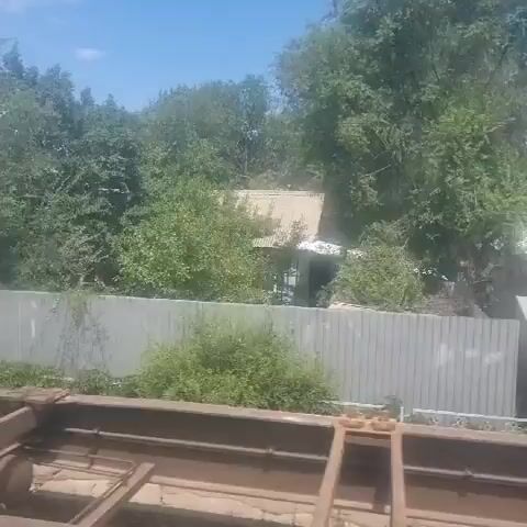 The video shows an outdoor scene under a clear blue sky with green trees and vegetation. In the foreground, there is a metallic structure, possibly part of a train car or industrial equipment, with rusty metal beams. The middle ground features several buildings. One building is a pale yellow, modular structure with a white, sloped roof and blue trim. It has a logo on the side and a red fire extinguisher mounted outside. Next to it is a smaller, white building with a blue trim and a white sloped roof. A dark minivan is parked in front of the yellow building, and a silver SUV is parked further down the street. Beyond these buildings, there are more structures, including a grey metal fence, smaller buildings with pitched roofs, and a white house with a green roof and a prominent window. There are also several cars visible in the distance. The scene appears to be a street or an industrial area with some residential elements. People are briefly visible near one of the buildings, and there is a utility pole with wires in the middle of the frame. The overall impression is of a somewhat rural or industrial setting on a sunny day.