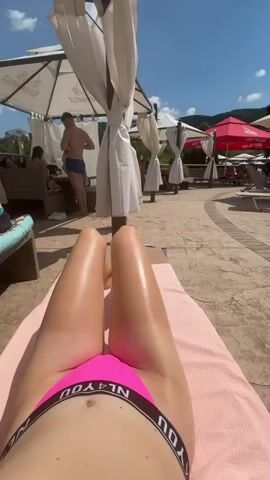 The video shows a resort pool area on a sunny day. The camera perspective is from a lounge chair, focusing on a person's legs in a pink bikini bottom. The background reveals rows of lounge chairs and umbrellas, with several people relaxing. A man in blue swim trunks stands near some of the covered seating areas. The ground is paved with stone, and lush green foliage and hills are visible in the distance under a bright blue sky with scattered clouds.