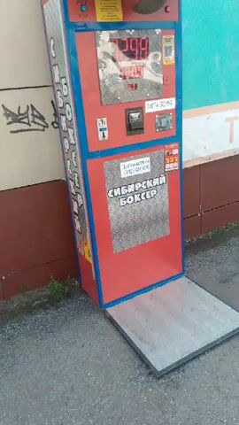 This is a video of a boxing game arcade machine, likely an outdoor installation, as indicated by the presence of gravel and what appears to be a building facade in the background. The machine is predominantly red with blue trim and features a digital display at the top, showing numbers, and a coin slot. There is also a target area where a player would hit a punch bag.

Visible text on the machine includes:
- "СИБИРСКИЙ БОКСЕР" (Siberian Boxer) which appears to be the name of the game.
- "1 ИГРА - 30 РУБЛЕЙ" (1 Game - 30 Rubles), indicating the cost to play.
- "ТЕХПОДДЕРЖКА +7923-501-22-72" (Technical Support +7923-501-22-72), providing a contact number.

The digital display at the top of the machine shows what appears to be a score or a countdown, with the numbers "44" and "500" visible at different points in the video. There are also some smaller text labels and icons, including what looks like a QR code for payment. The overall condition of the machine suggests it is functional and in use. The lighting suggests it is daytime, and there are no overt indications of weather conditions. The scene is static, with the camera panning slightly, focusing on the details of the machine.
