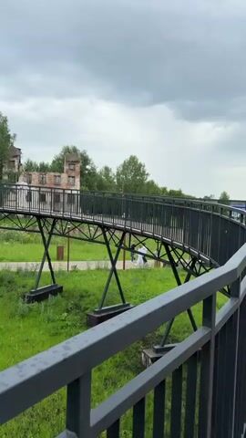 A slightly elevated, curved metal pedestrian bridge with black railings crosses a grassy area. In the background, the dilapidated brick ruins of a building stand amongst trees. Several people, including a group in white clothing, are walking on the pathway below the bridge. The sky is overcast with gray clouds.