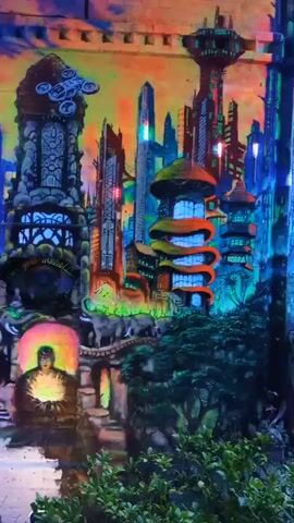 A video pans across a vibrant, detailed mural painted on a brick wall. The artwork depicts a futuristic cityscape with towering, intricate buildings, some with mushroom-shaped rooftops and others resembling ornate spires. A drone hovers near the top of a tall, gothic-style tower on the left. In the lower left section, a figure sits in a meditative pose, glowing with light, surrounded by what appear to be elephants and ornate archways. Further to the left, a skull with tree branches growing from its eye sockets is visible. The mural is illuminated with neon and fluorescent colors, creating a psychedelic and otherworldly atmosphere. The background transitions from a warm, orange and yellow sky to a deep blue and purple night sky. Green foliage frames the bottom of the scene. The camera moves slowly from left to right, revealing more of the mural's complexity.