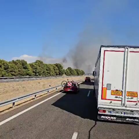 THE VIDEO SHOWS A HIGHWAY WITH SEVERAL TRUCKS AND CARS PARKED. A LARGE PLUME OF SMOKE IS VISIBLE IN THE BACKGROUND, INDICATING A FIRE. ONE CAR HAS A BICYCLE MOUNTED ON ITS ROOF. THE SKY IS CLEAR AND BLUE. THE SCENE APPEARS TO BE A TRAFFIC DELAY DUE TO THE FIRE.