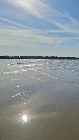 The video shows a beach scene. The sun is shining brightly and reflecting off the wet sand and shallow water. The sky is blue with some white clouds. There is a line of buildings or trees in the distance. The camera is stationary and pointed towards the horizon. The overall mood is peaceful and serene.