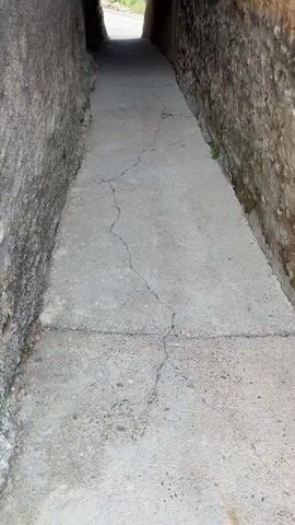 A narrow, outdoor stone alleyway with a concrete path leads to a bright opening. The walls of the alley are made of rough, aged stone, with some sections appearing to be plastered or crumbling. On the left side, there is a wall with a grey textured surface and a dark barred window. The right wall is primarily exposed stone. The concrete path has visible cracks. The opening at the end of the alley reveals greenery and a glimpse of what appears to be a road. The perspective is from a low angle, looking forward through the alley.