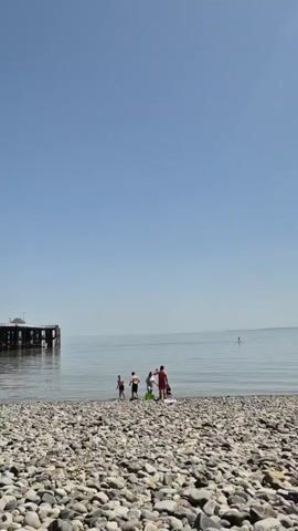 The video shows a group of people enjoying a sunny day at a rocky beach, with a pier extending into the calm sea. The beach is covered with smooth, gray and white stones. In the distance, a pier structure with white buildings on it juts out into the water. A few individuals are standing near the water's edge, while others are positioned closer to the pier. The sky is clear and blue.