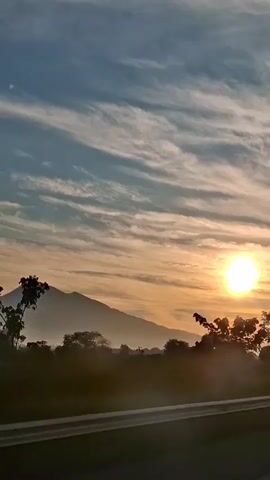 The video shows a sunrise over a mountainous landscape. The sun is a bright orb in the sky, casting a warm glow over the scene. Wispy clouds streak across the blue sky, catching the light. In the foreground, trees and vegetation are silhouetted against the rising sun. A road or highway is visible in the bottom of the frame, suggesting the video was taken from a moving vehicle. The overall impression is one of peace and natural beauty.