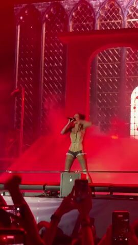 A female performer on a stage is seen wearing a corset and shorts with thigh-high stockings. The stage features large, arched windows and is lit with red and yellow lights. The performer is holding a microphone and singing. Below the stage, a crowd of people are holding up their phones to record the performance. The audience is visible in the foreground, with many hands holding up smartphones. The overall atmosphere appears to be a concert or live performance.